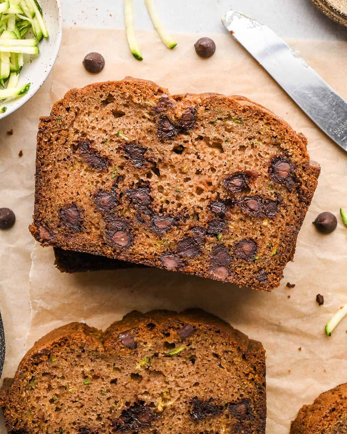 3 slices of chocolate chip zucchini bread and the loaf on parchment paper with shredded zucchini and chocolate chips nearby