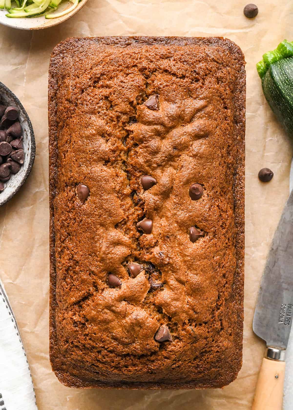 a whole loaf of chocolate chip zucchini bread on parchment paper with zucchini and a bowl of chocolate chips 