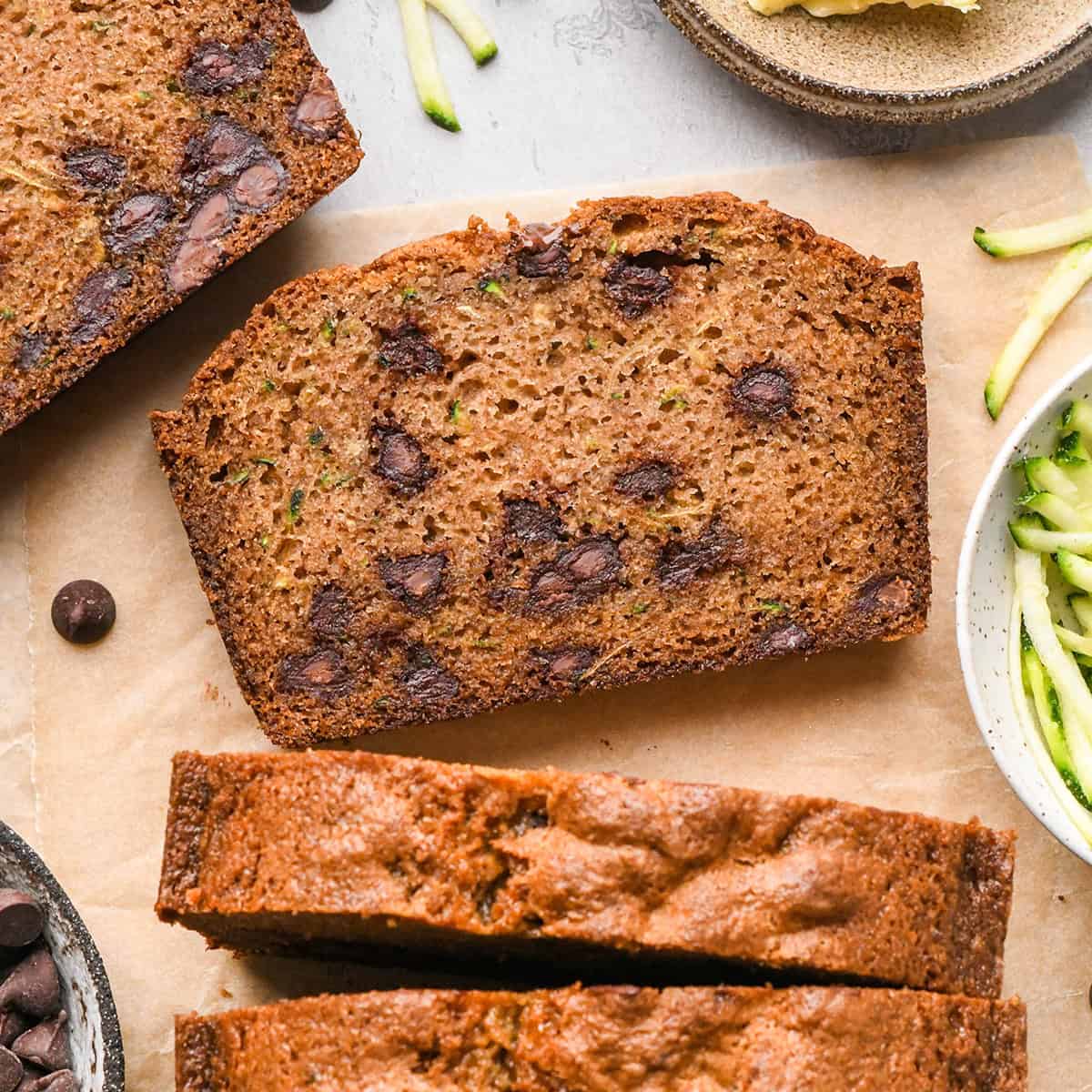 two slices of chocolate chip zucchini bread and the loaf on parchment paper with shredded zucchini and chocolate chips nearby