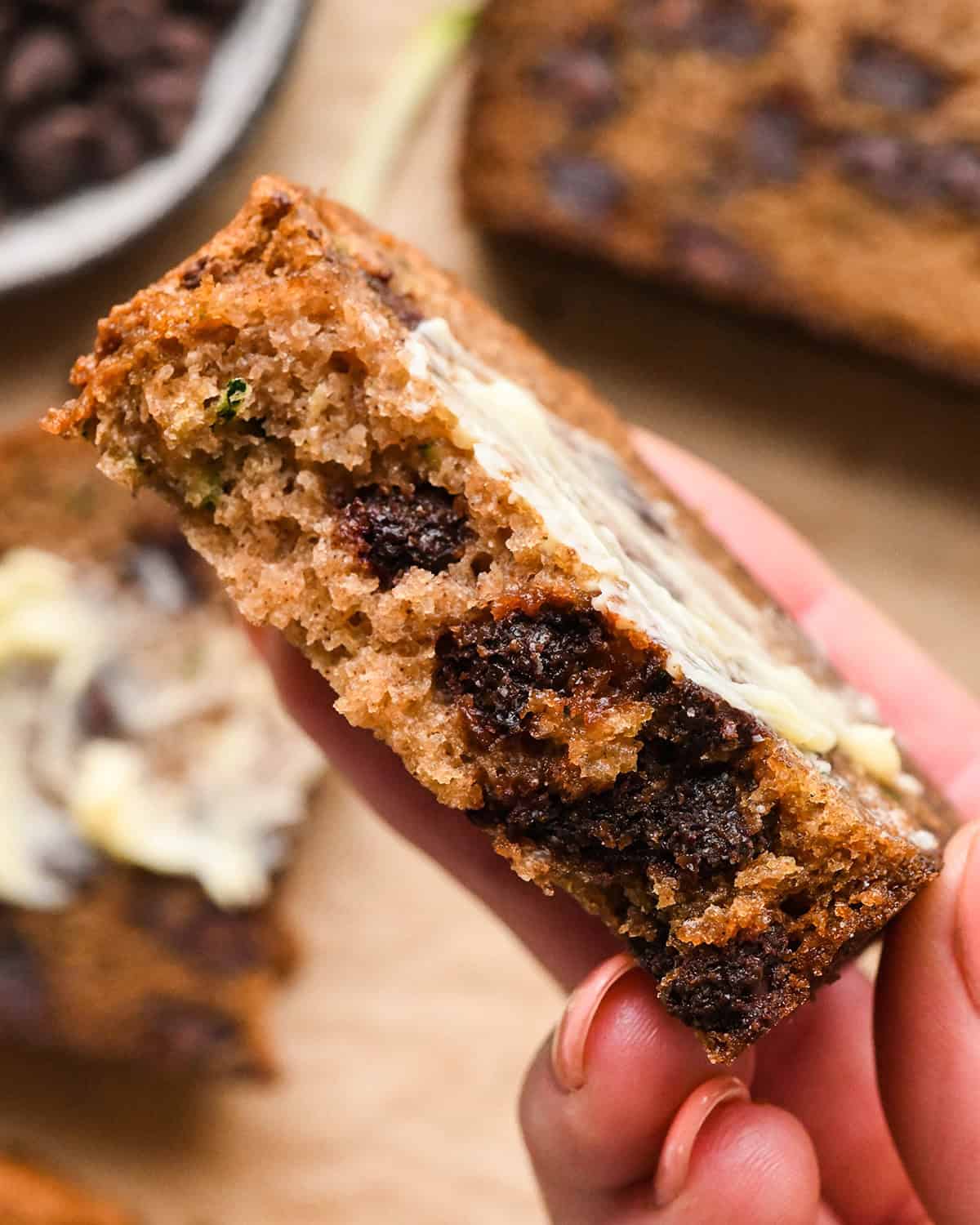 a hand holding a slice of Chocolate Chip Zucchini Bread with a bite taken out of it and butter spread on top 