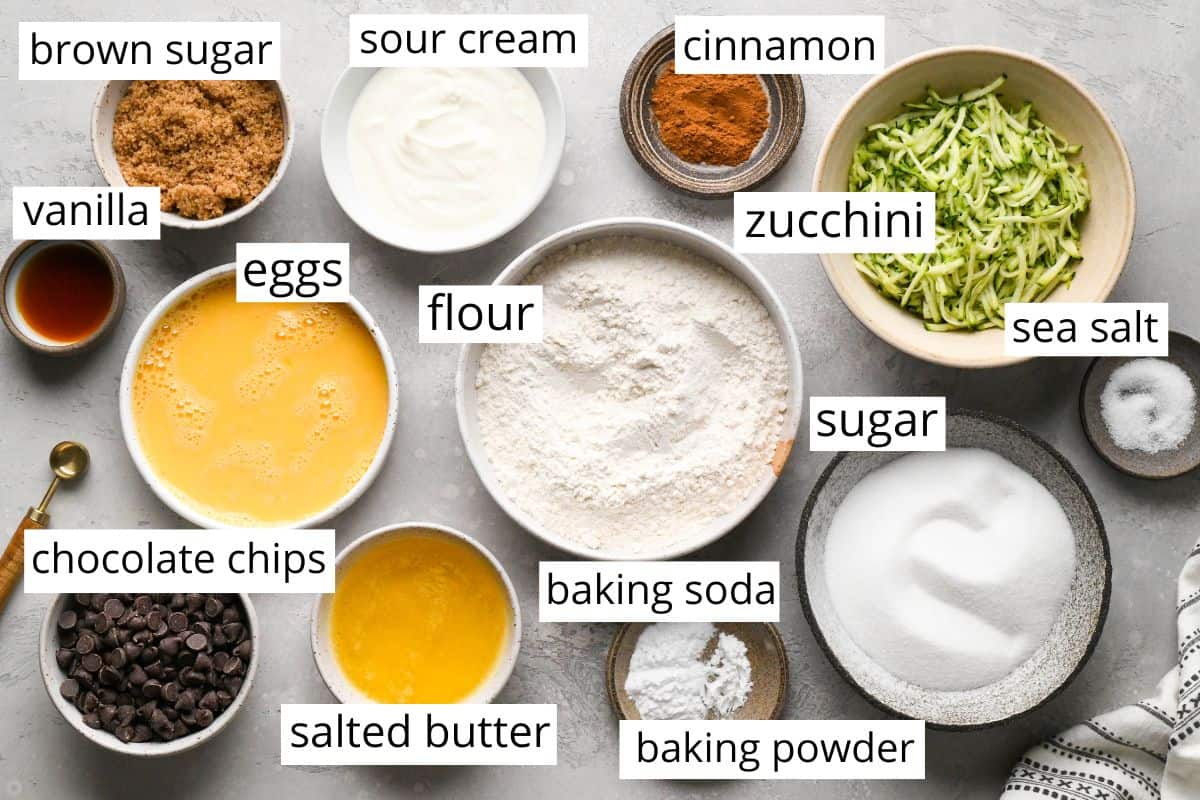 overhead photo of the labeled ingredients in this Chocolate Chip Zucchini Bread recipe in bowls like flour, zucchini, sugar, butter, eggs, brown sugar, sour cream, and chocolate chips