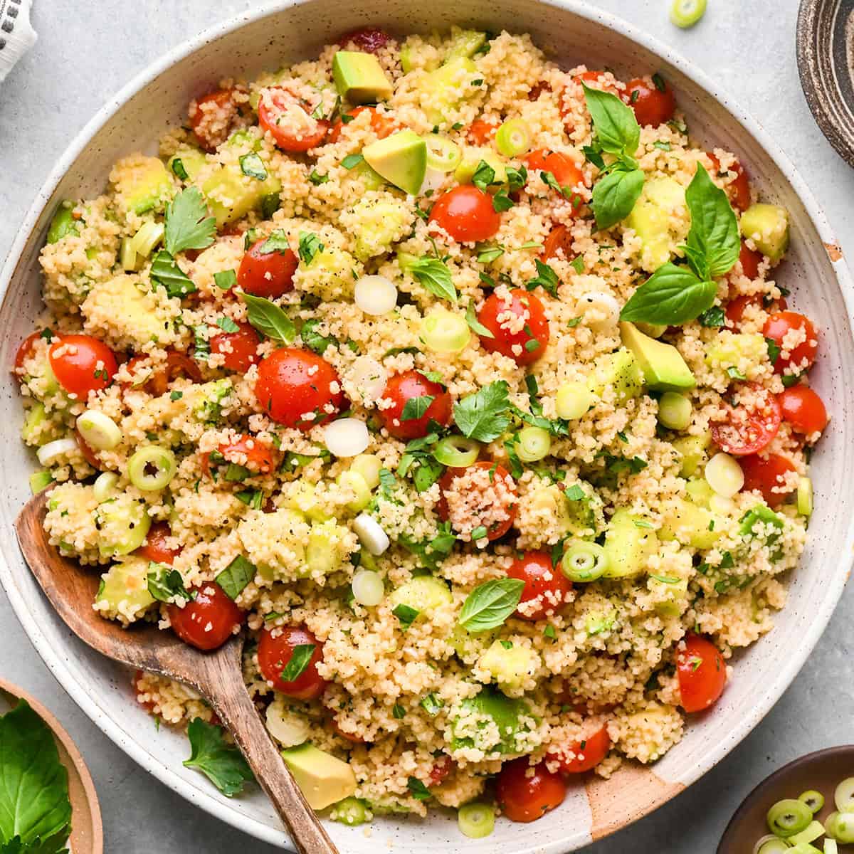 couscous salad with cherry tomatoes, avocado, green onions, and fresh herbs in a large bowl with a wooden spoon on a gray surface