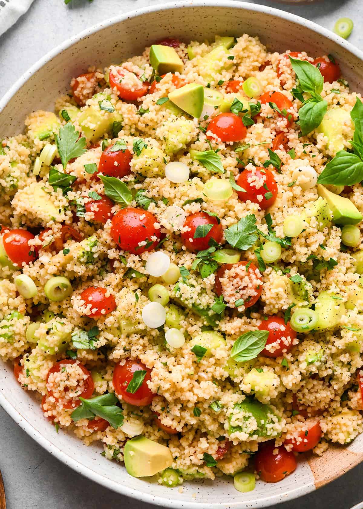 couscous salad with cherry tomatoes, avocado, green onions, and fresh herbs in a large bowl on a gray surface
