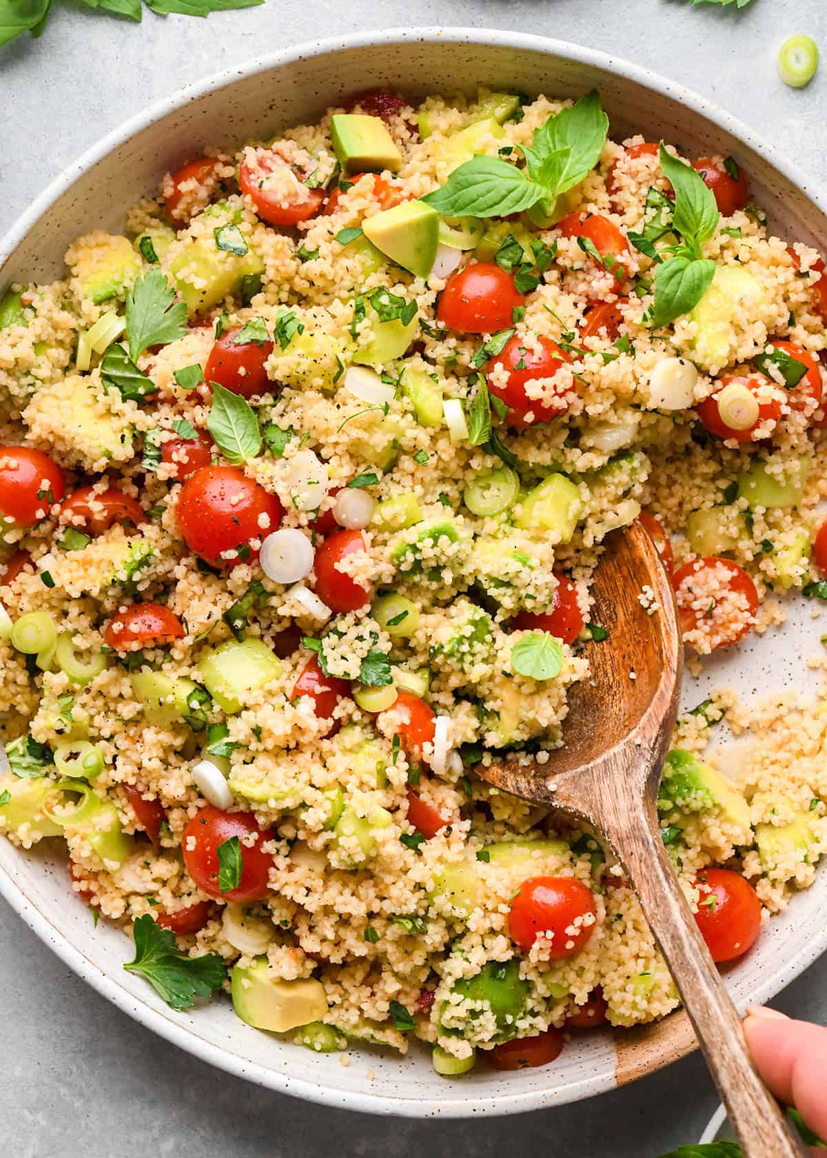 a wooden spoon taking a scoop of couscous salad containing cherry tomatoes, avocado, green onions, and fresh herbs in a large bowl