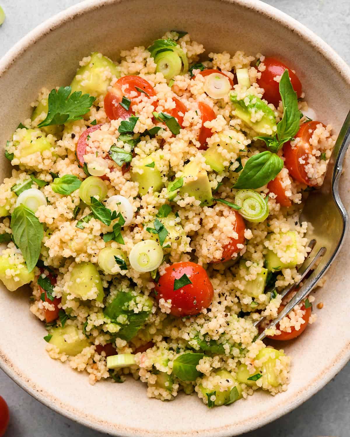 couscous salad with cherry tomatoes, avocado, green onions, and fresh herbs in a small bowl with a fork