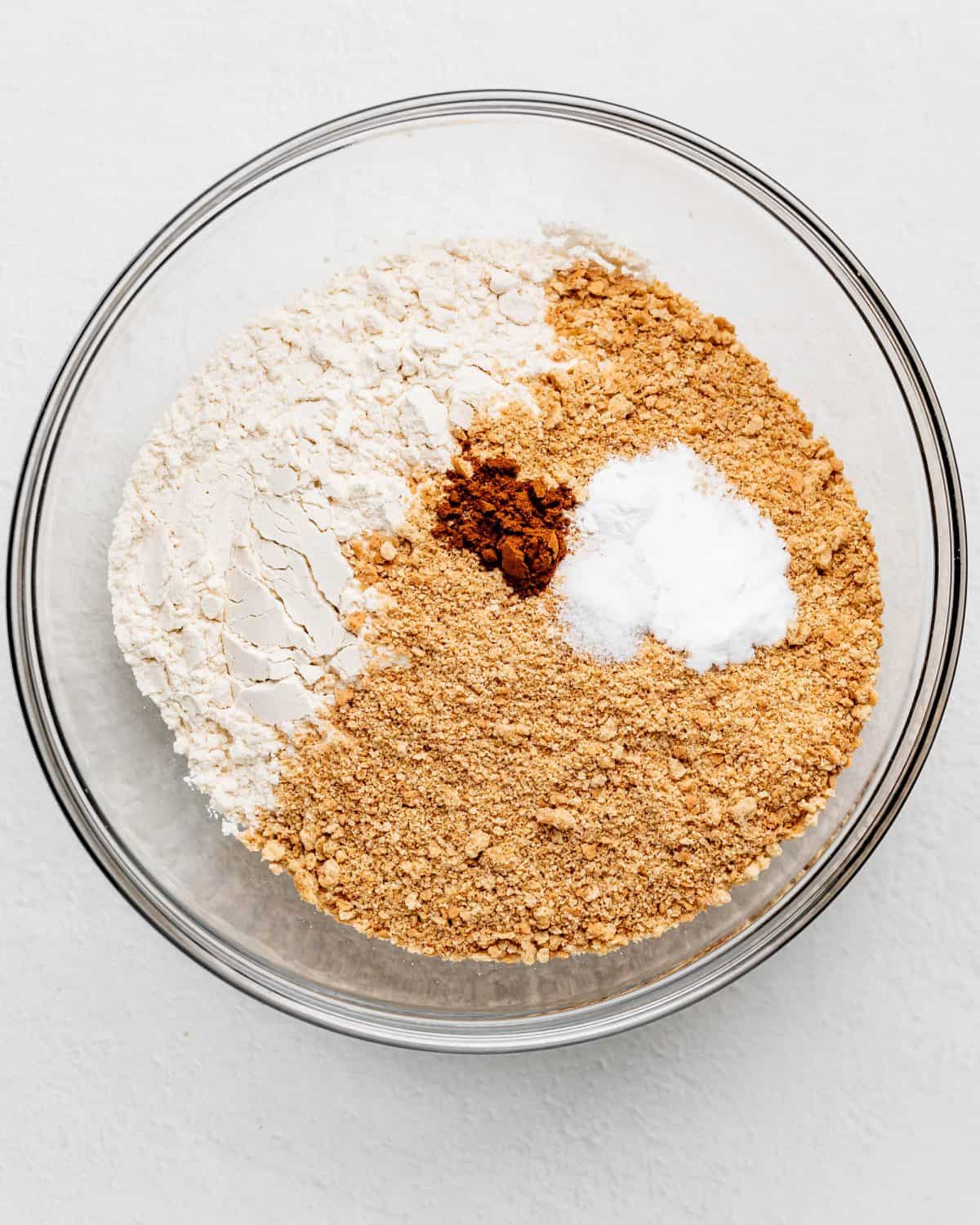 making s'mores cookies recipe - dry ingredients including flour, graham cracker crumbs, salt, baking soda, baking powder and cinnamon in a glass mixing bowl on a white surface before mixing
