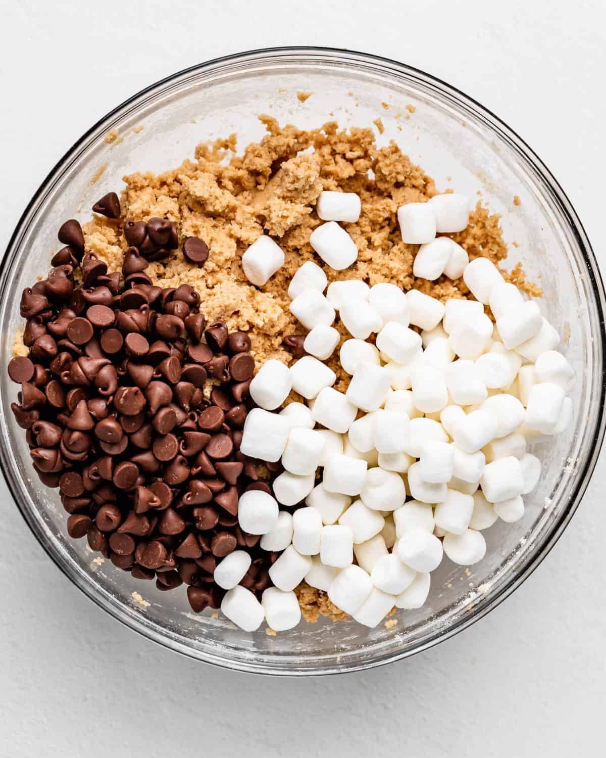 making s'mores cookies recipe - chocolate chips and mini marshmallows added to the dough before mixing in a glass bowl on a white surface