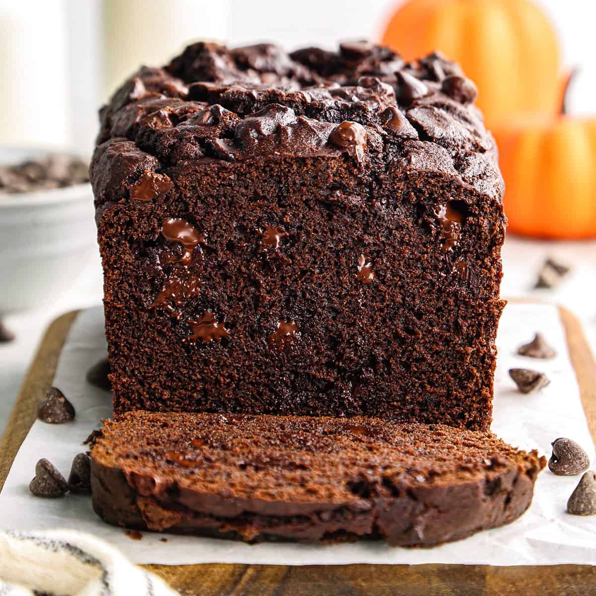a loaf of chocolate pumpkin bread with melty chocolate chips with a slice cut out of it on a wooden board with parchment paper and chocolate chips. There's a bowl of chocolate chips and pumpkins in the background