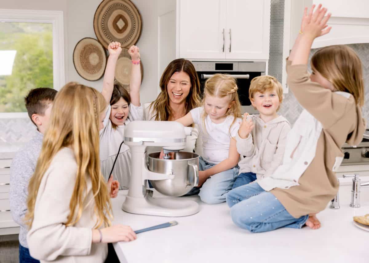 a mom and her 6 kids gathered around a standing mixer laughing