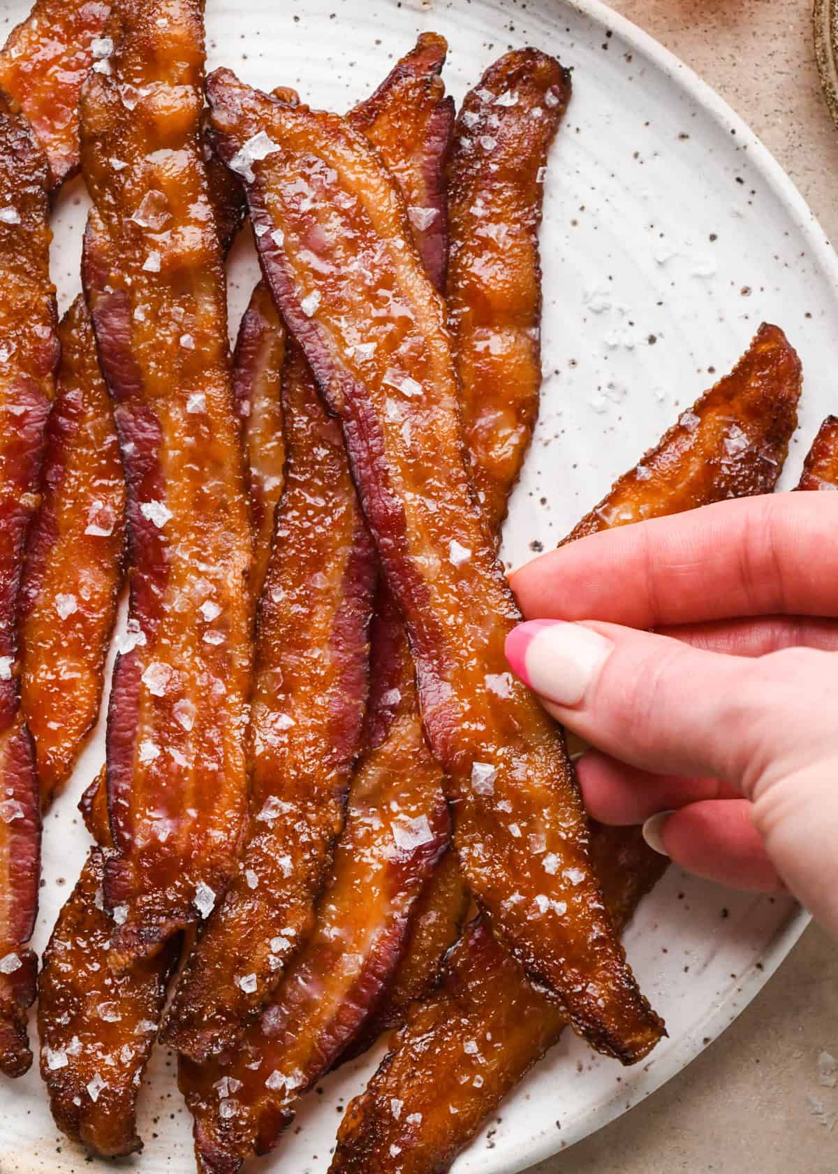 Crispy maple-glazed bacon strips topped with sea salt and served on a white speckled plate - a person is holding one piece of bacon
