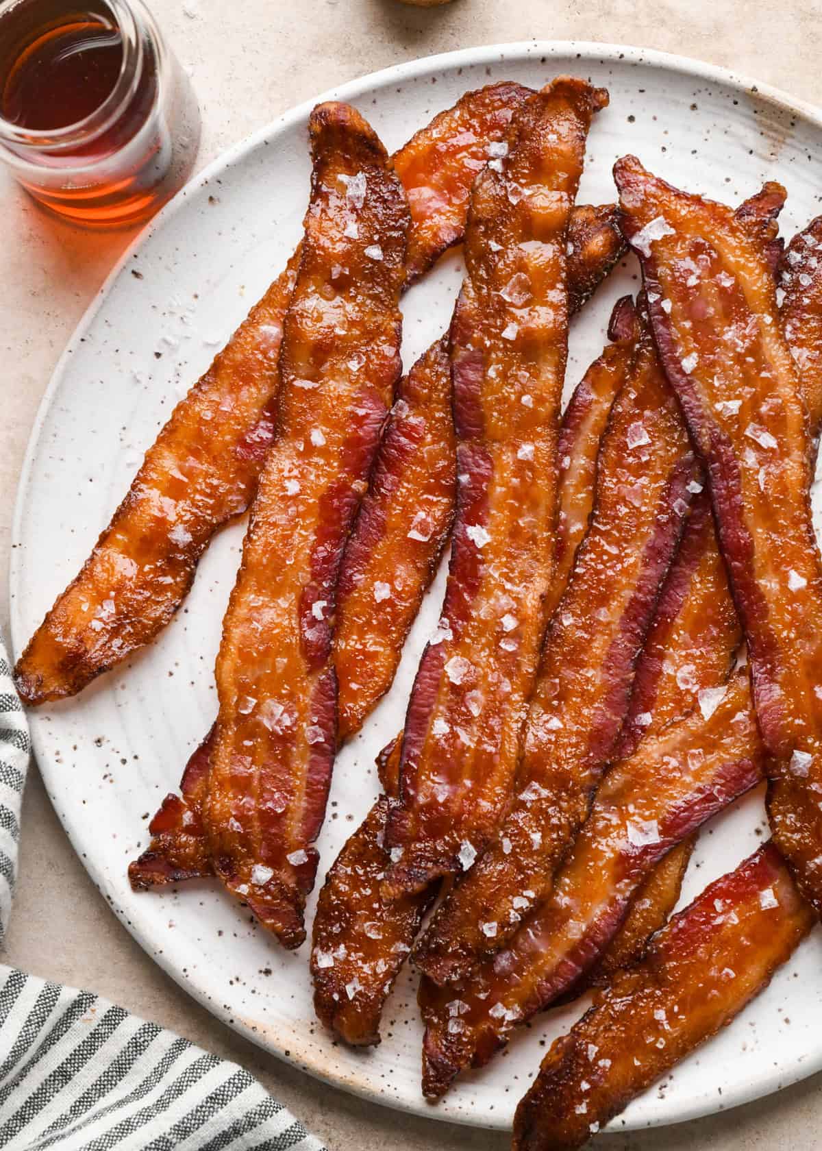 Crispy maple-glazed bacon strips topped with flaky sea salt on a white speckled plate served next to a clear jar of maple syrup
