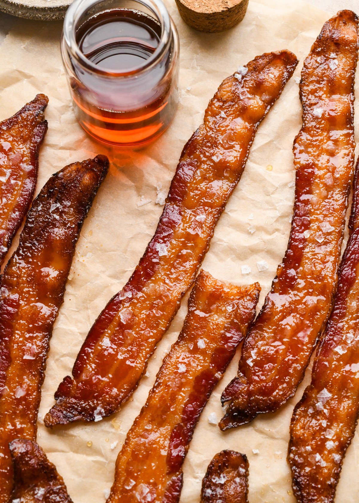Crispy maple-glazed bacon strips topped with flaky sea salt on parchment paper next to a clear jar of maple syrup