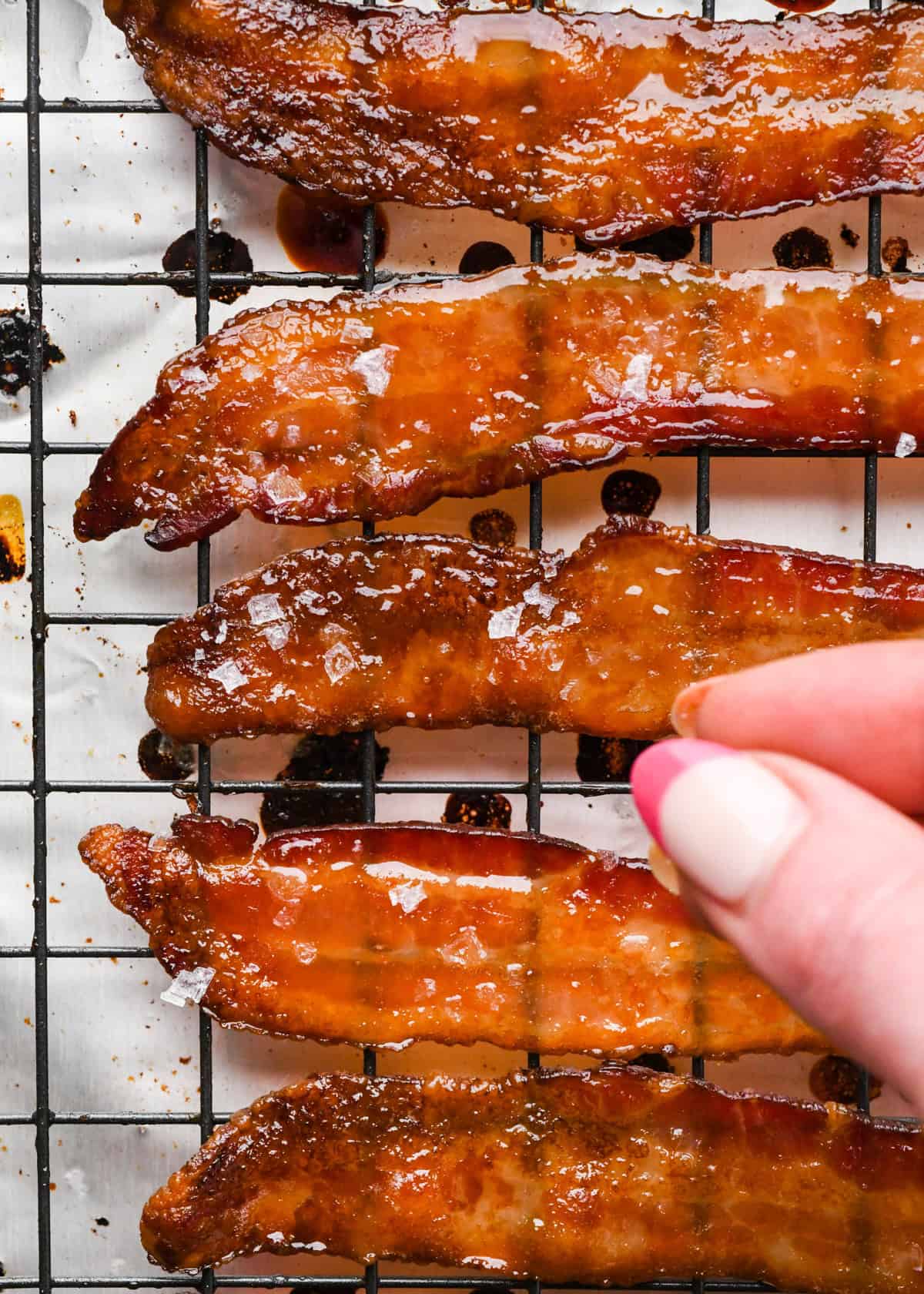 Overhead photo of crispy maple-glazed bacon strips after they've been baked in the oven, being topped with flaky sea salt