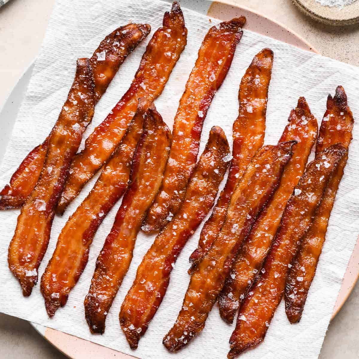 Overhead photo of crispy maple-glazed bacon strips topped with flaky sea salt and sitting on a paper-towel-covered plate