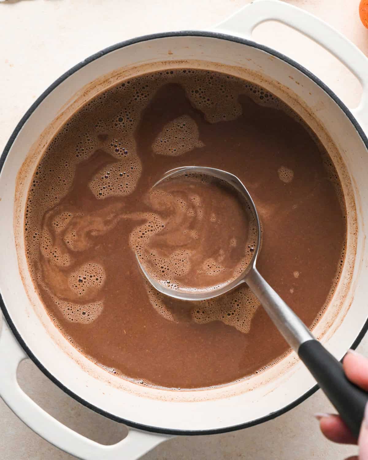 a ladle taking a scoop of pumpkin spice hot chocolate out of a white pot 