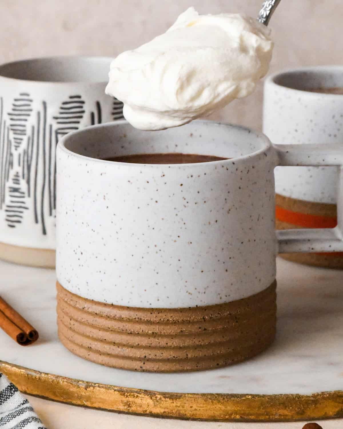 a spoon of whipped cream being put into a ceramic mug of pumpkin spice hot chocolate with two other mugs and a cinnamon stick in the background