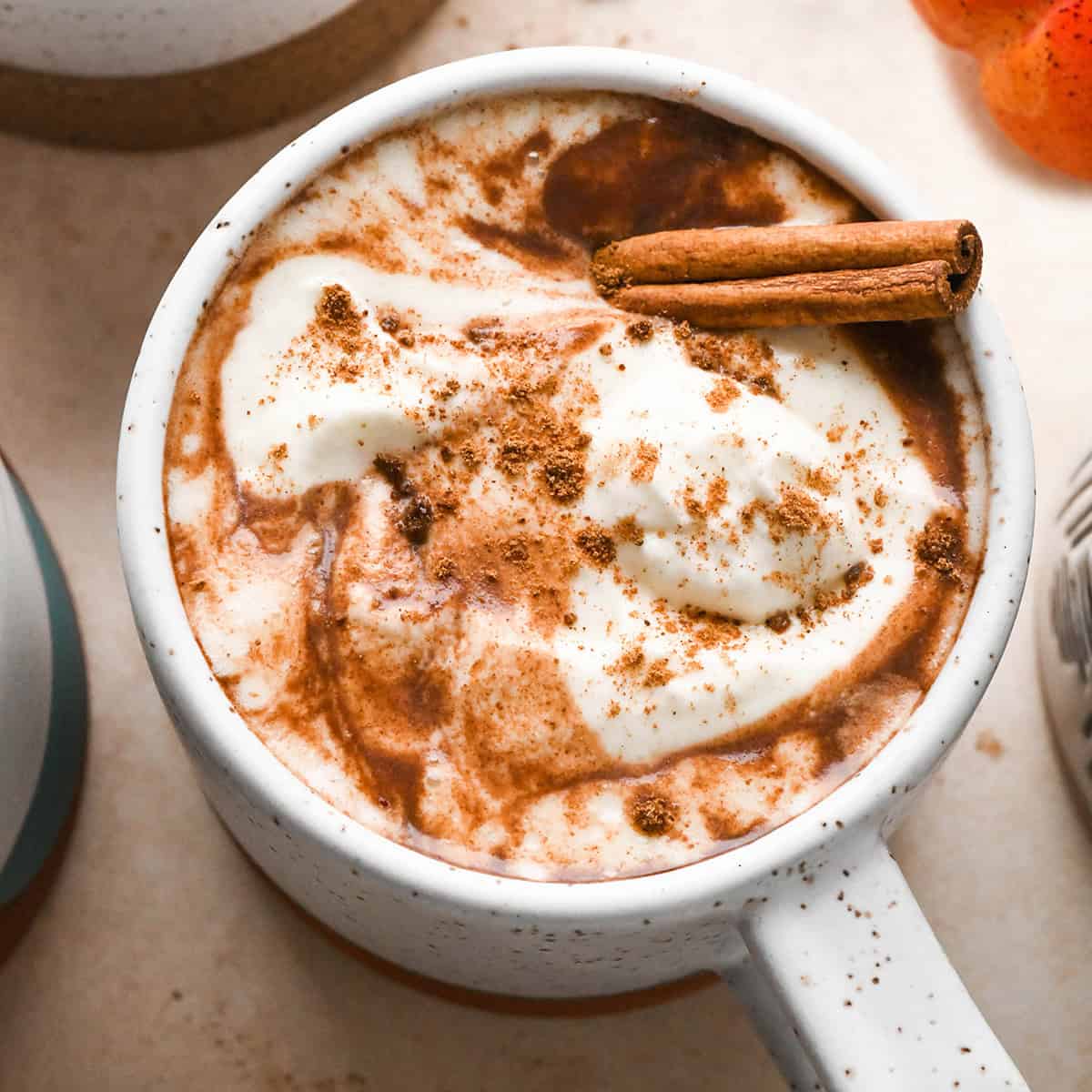 a mug filled with pumpkin hot chocolate, topped with whipped cream and pumpkin spice, and garnished with a cinnamon stick. there's a pumpkin and another mug in the background