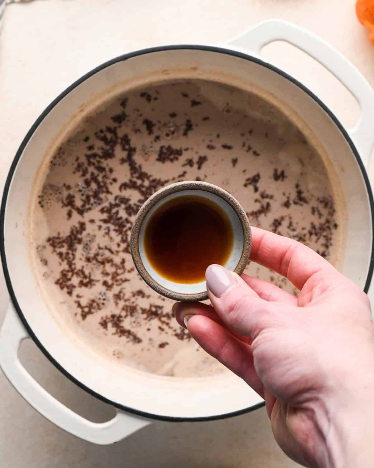 a hand holding a a small bowl of vanilla extract over a white pot making pumpkin spice hot chocolate