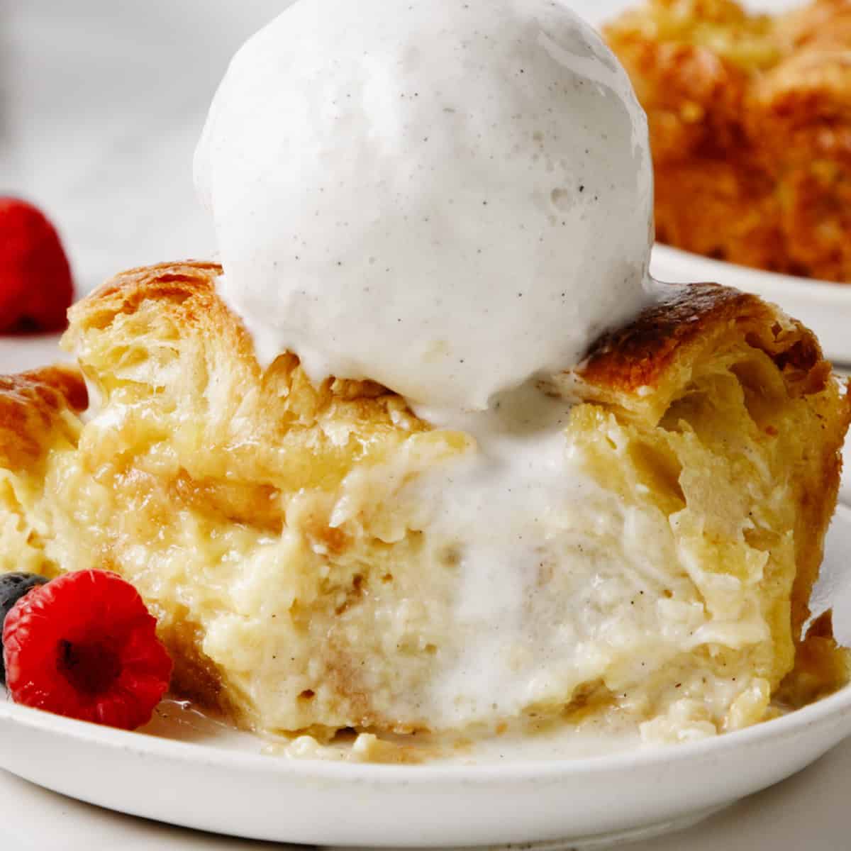 a piece of croissant bread pudding on a plate topped with vanilla ice cream. A bite is taken out of it and berries are on the plate