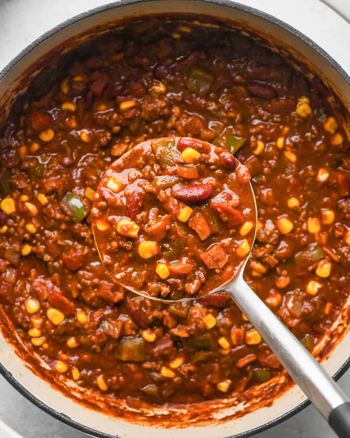 Making bacon chili: up-close photo of completed chili in a white pot, being scooped up with a metal ladle.