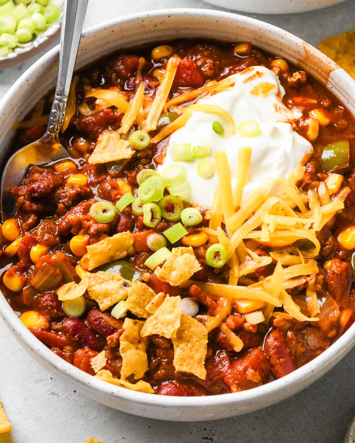 A white bowl of hearty bacon chili topped with sour cream, shredded cheddar cheese, sliced green onions, and crushed tortilla chips, with a spoon resting in the bowl on a light gray surface.
