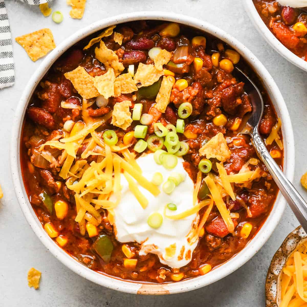 A white bowl of hearty bacon chili topped with sour cream, shredded cheddar cheese, sliced green onions, and crushed tortilla chips, with a spoon resting in the bowl on a light gray surface.