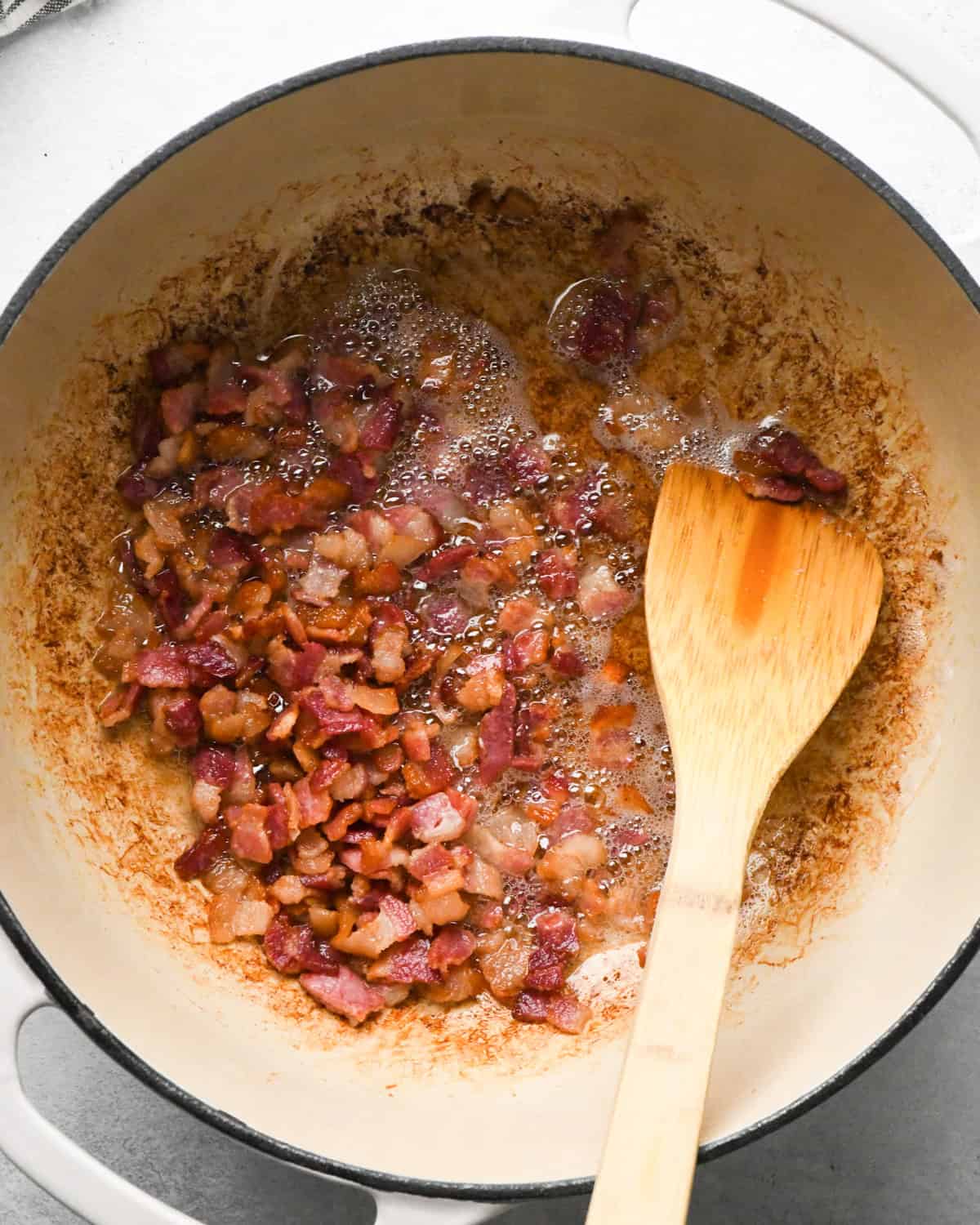 Making bacon chili: cooking bacon in a white skillet, stirring with a wooden spoon.