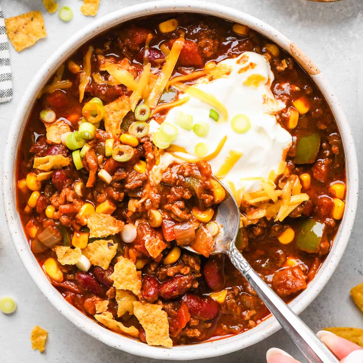 Making bacon chili: up-close photo of completed chili in a white speckled bowl, with sour cream, cheese, and green onions on top and being eaten with a metal spoon.