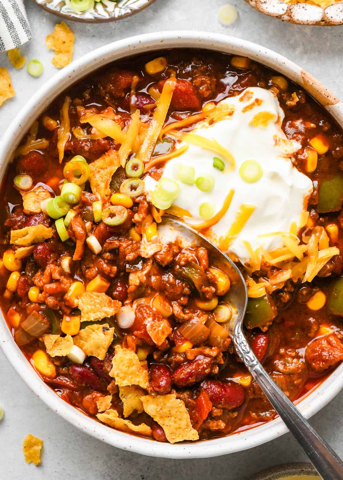 Making bacon chili: up-close photo of completed chili in a white speckled bowl, with sour cream, cheese, and green onions on top and being eaten with a metal spoon.