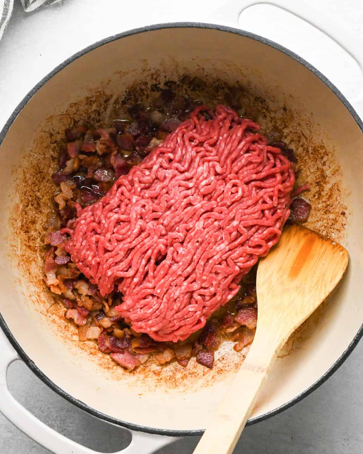 Making bacon chili: cooking bacon and ground beef in a white skillet, stirring with a wooden spoon.