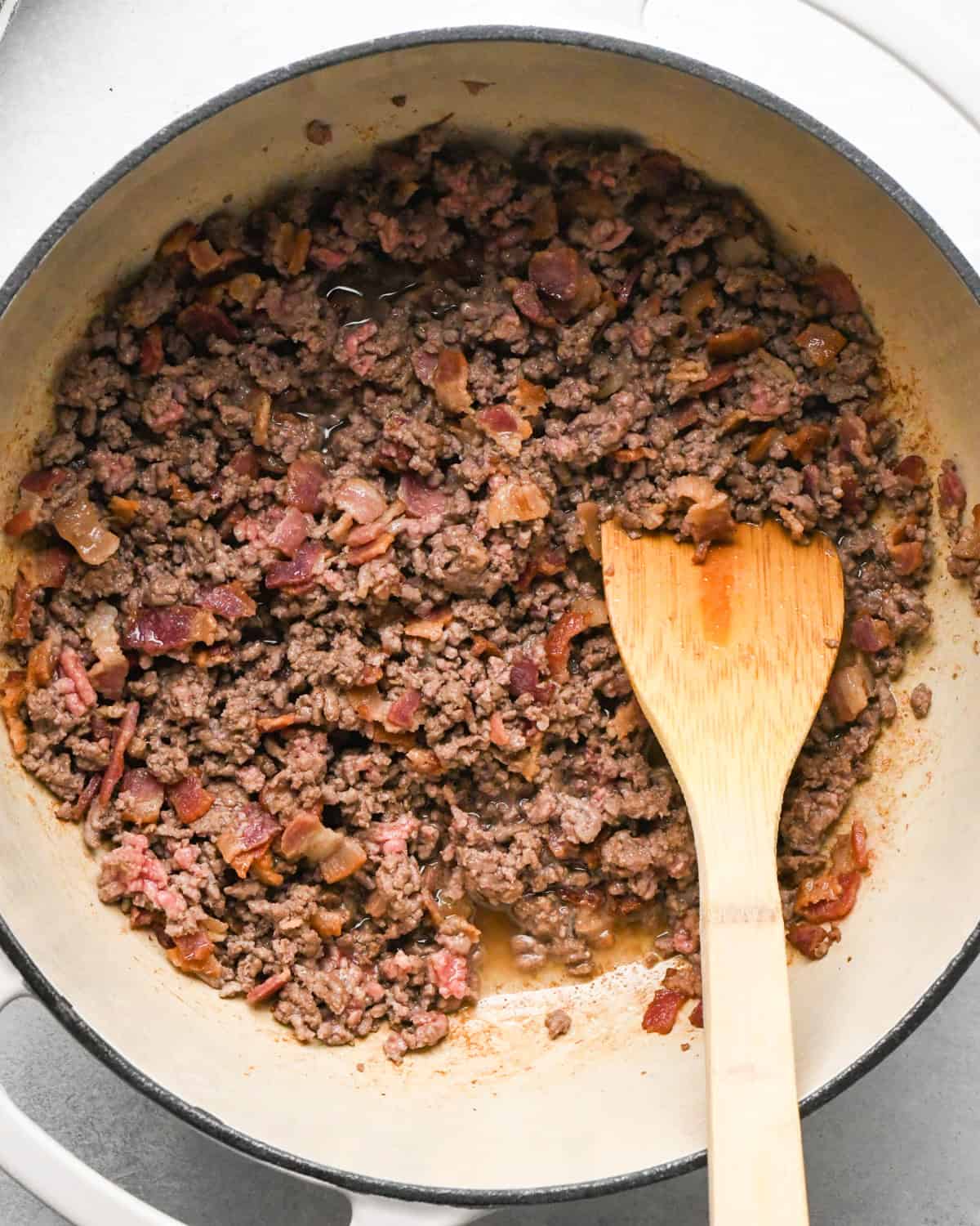 Making bacon chili: cooking bacon and ground beef in a white skillet, stirring with a wooden spoon.