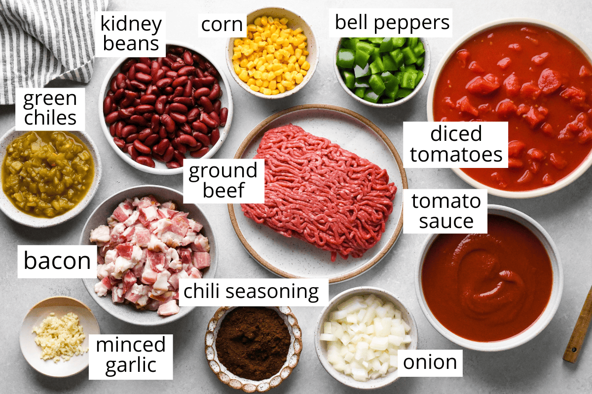 Overhead view of ingredients for bacon chili, including ground beef, chopped bacon, kidney beans, diced tomatoes, tomato sauce, corn, bell peppers, green chiles, onion, minced garlic, and chili seasoning arranged in bowls on a gray surface.
