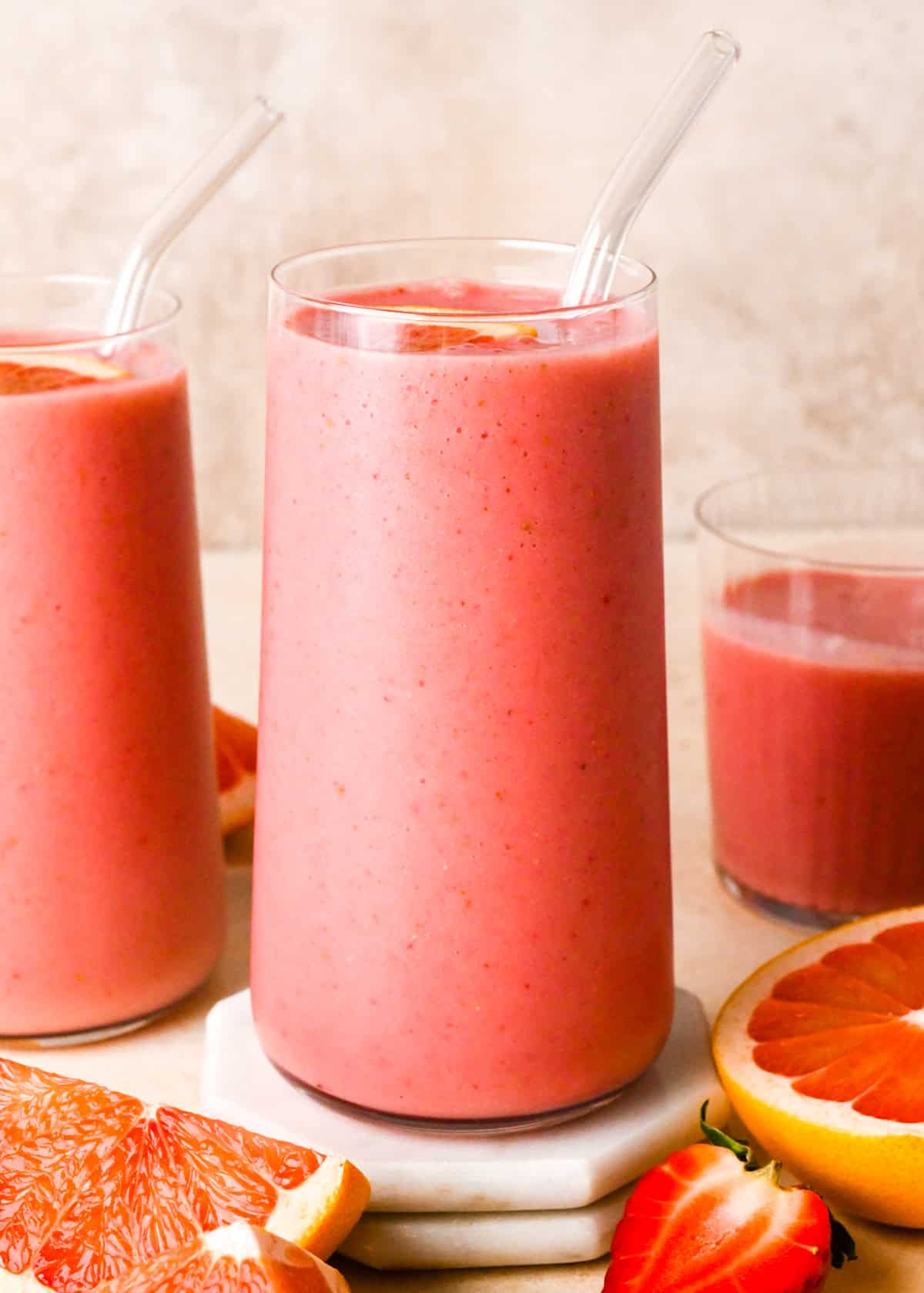 A creamy pink grapefruit smoothie served in a clear glass with a glass straw, topped with a wedge of fresh grapefruit. Other glasses of the same smoothie are visible in the background on a light surface.
