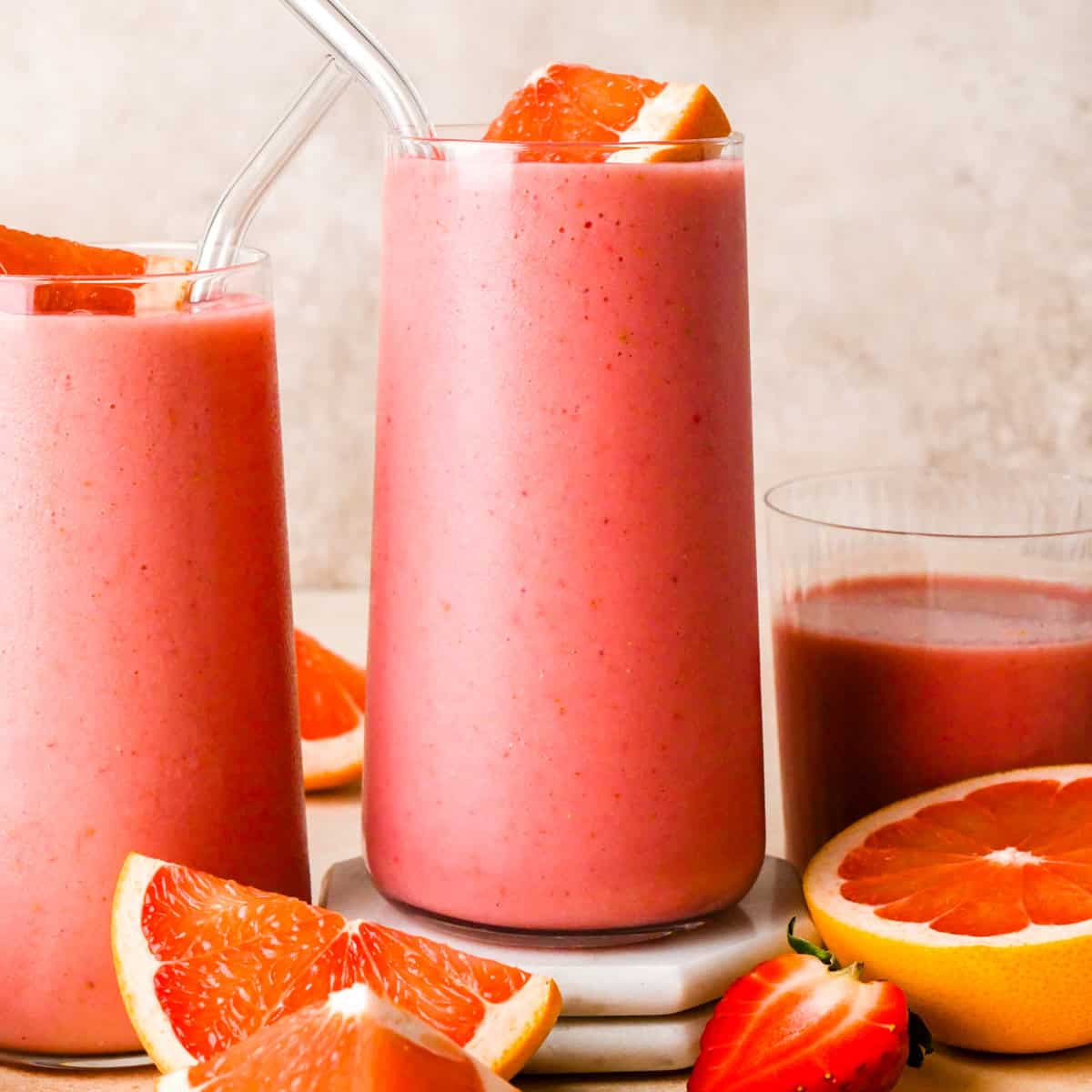 Close up photo of a grapefruit smoothie in a clear glass with a clear straw and topped with a slice of fresh grapefruit. Other glasses and slices of fruit are surrounding it on a light surface.