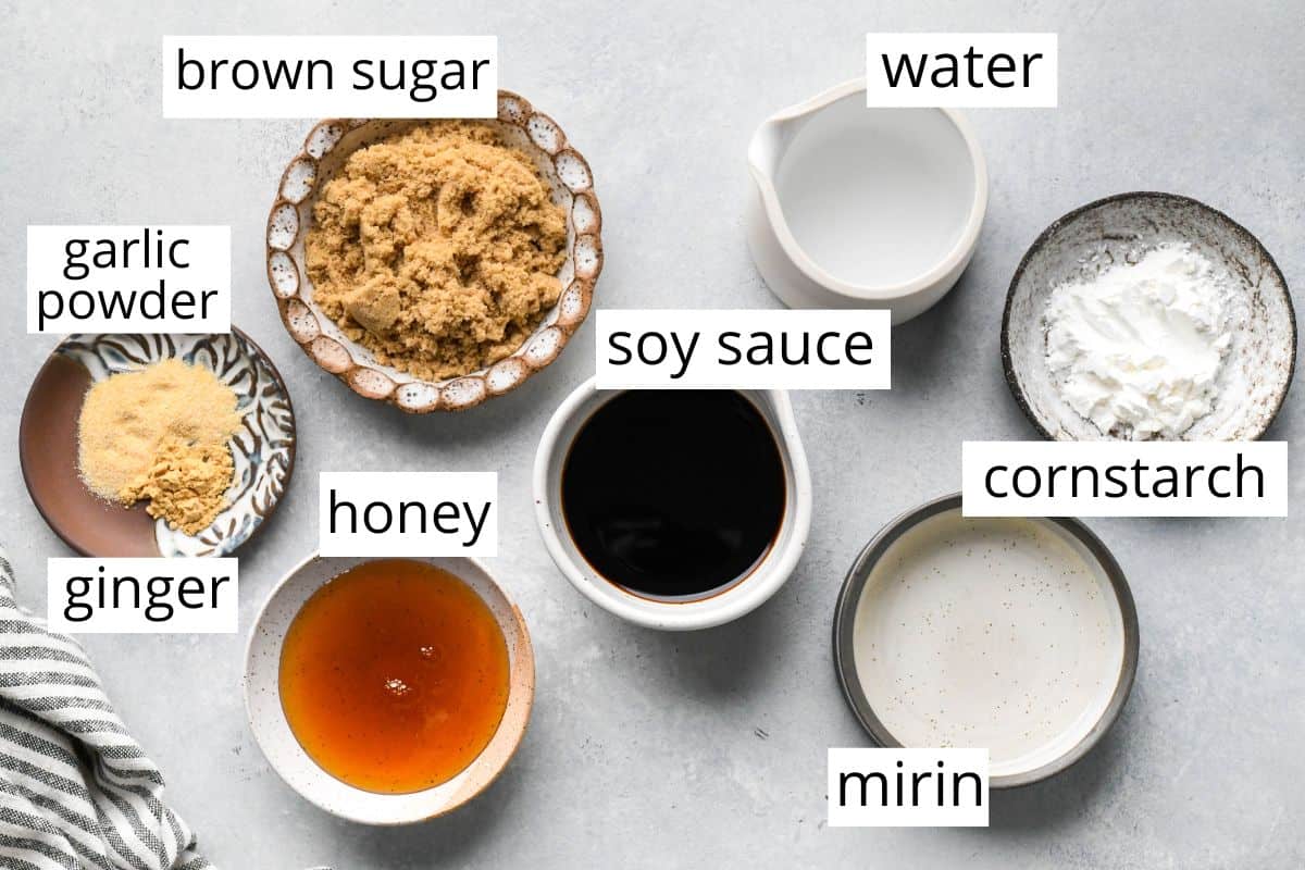 Top-down view of labeled ingredients for homemade teriyaki sauce, including brown sugar, soy sauce, water, cornstarch, mirin, honey, ginger, and garlic powder, arranged neatly on a light countertop.