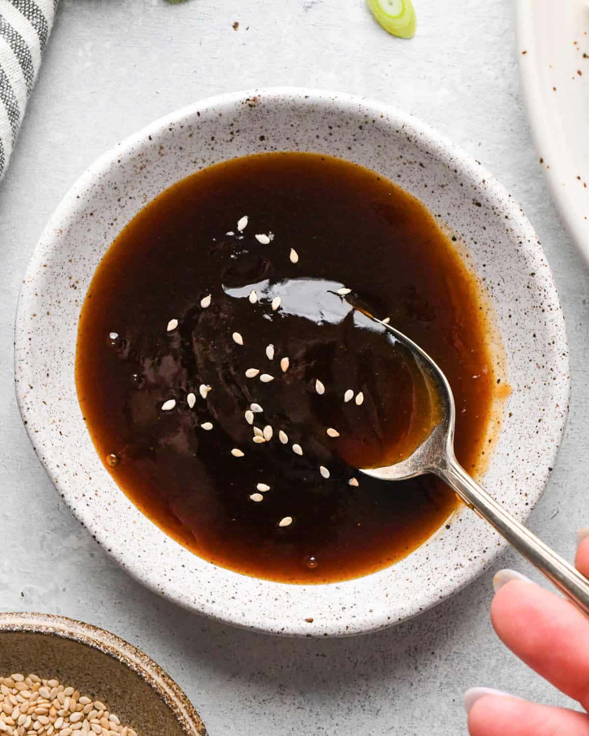 A small bowl of thick homemade teriyaki sauce topped with sesame seeds, with a spoon resting in the sauce.