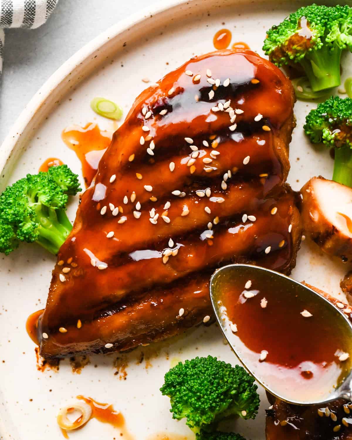 A plate of sliced chicken breast drizzled with glossy homemade teriyaki sauce, topped with sesame seeds and green onions, served with white rice and steamed broccoli.