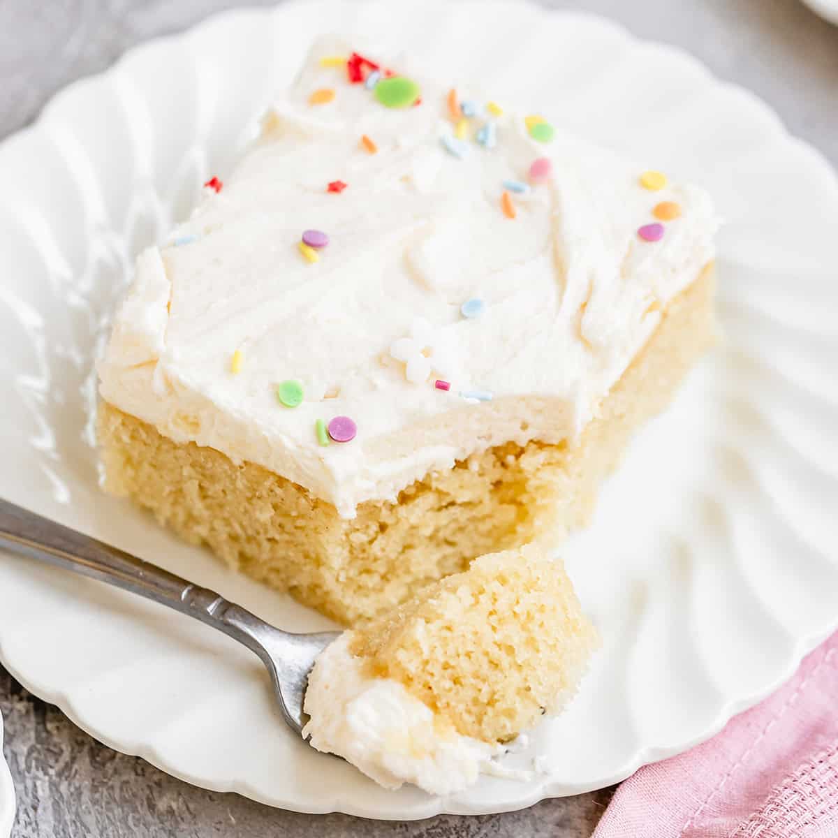 a fork taking a bite of a piece of Vanilla Sheet Cake with frosting and sprinkles on a plate
