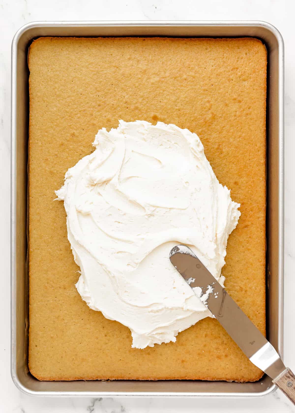 spreading vanilla frosting over the cake in a sheet pan