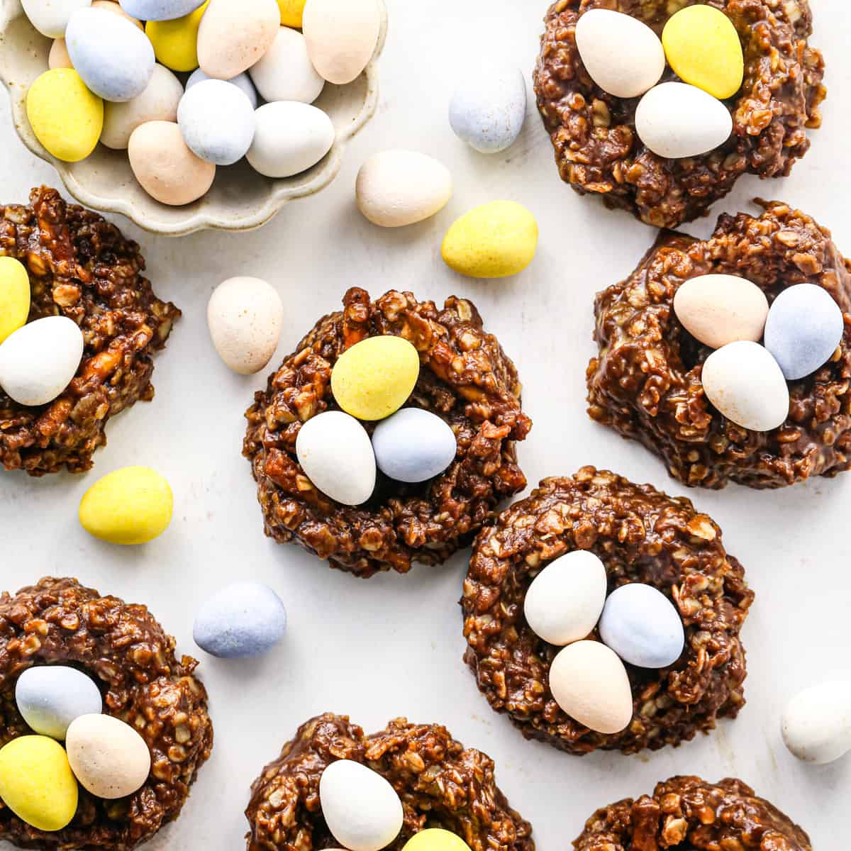 Overhead photo of chocolate bird’s nest cookies made with a homemade chocolate mixture, shaped into nests and filled with pastel candy eggs on a white surface.