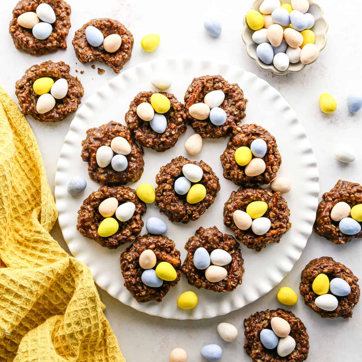 Overhead photo of chocolate bird’s nest cookies arranged on a white plate. The cookies are shaped into nests and filled with pastel candy eggs. The plate is surrounded by additional cookies, candy eggs, and a yellow tea towel on a white surface.