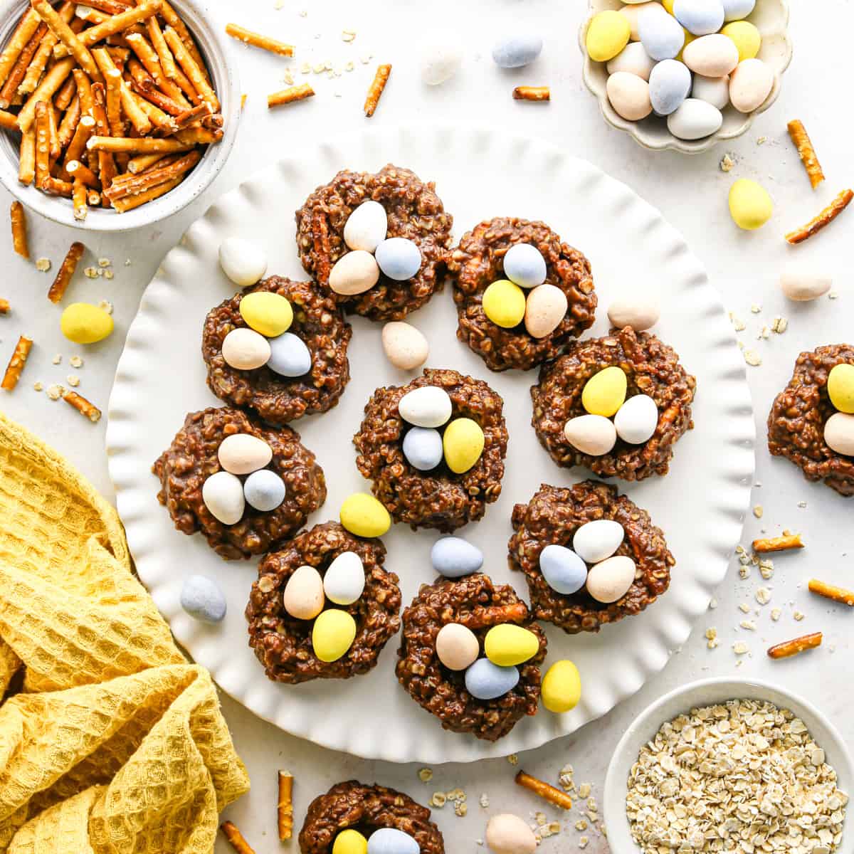 Overhead photo of chocolate bird’s nest cookies arranged on a white plate. The cookies are shaped into nests and filled with pastel candy eggs. The plate is surrounded by additional cookies, candy eggs, and a yellow tea towel on a white surface.