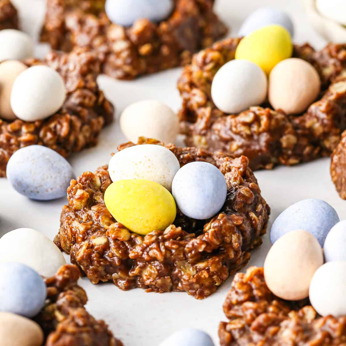 Close-up of chocolate bird’s nest cookies made with a homemade chocolate mixture, shaped into nests and filled with pastel candy eggs on a white surface.