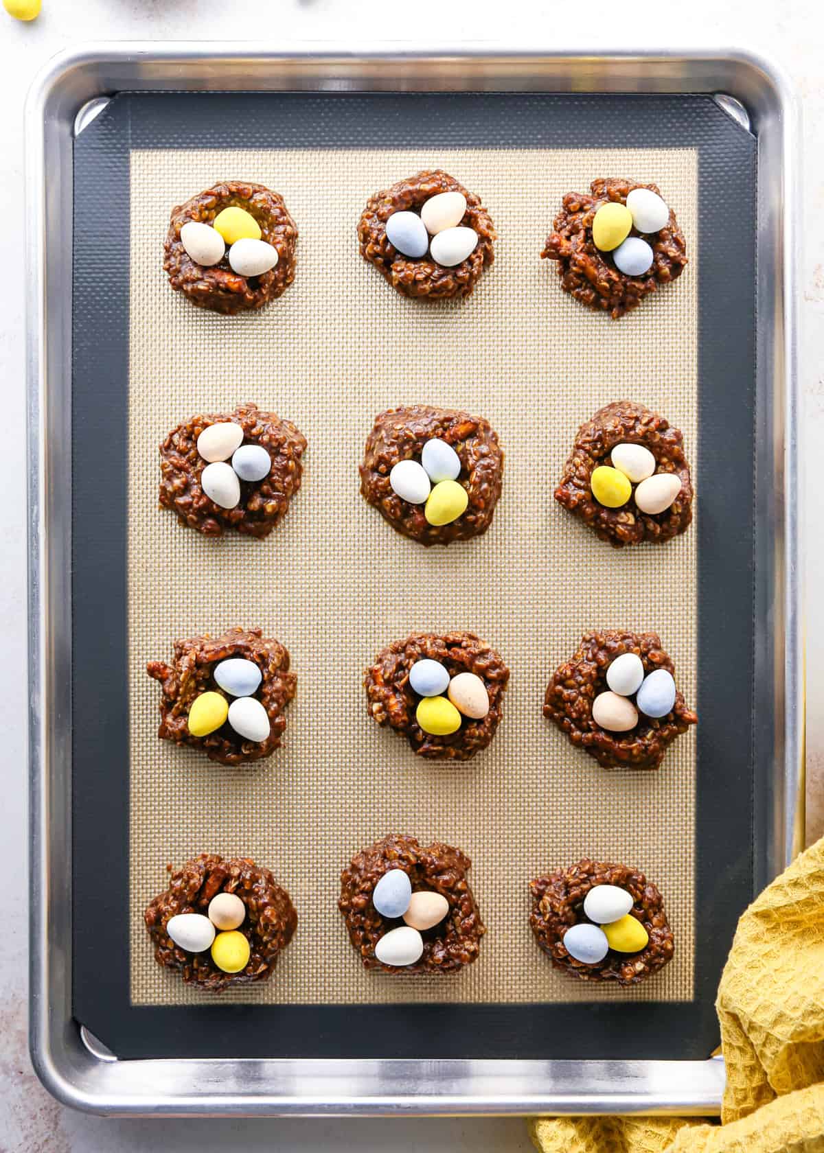 Making bird's nest cookies: overhead photo of 12 balls of cookie dough on a metal baking sheet. Each cookie dough ball has been topped with 3 pastel candy eggs.