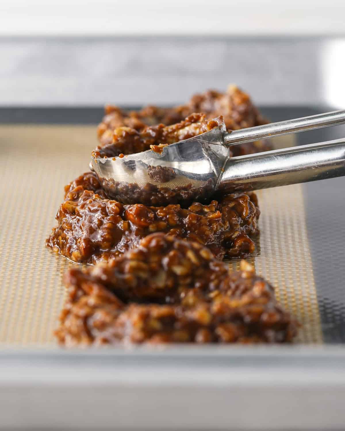 Making bird's nest cookies: close-up photo of a ball of cookie dough scooped onto a metal baking sheet and being pressed down with a cookie dough scoop.