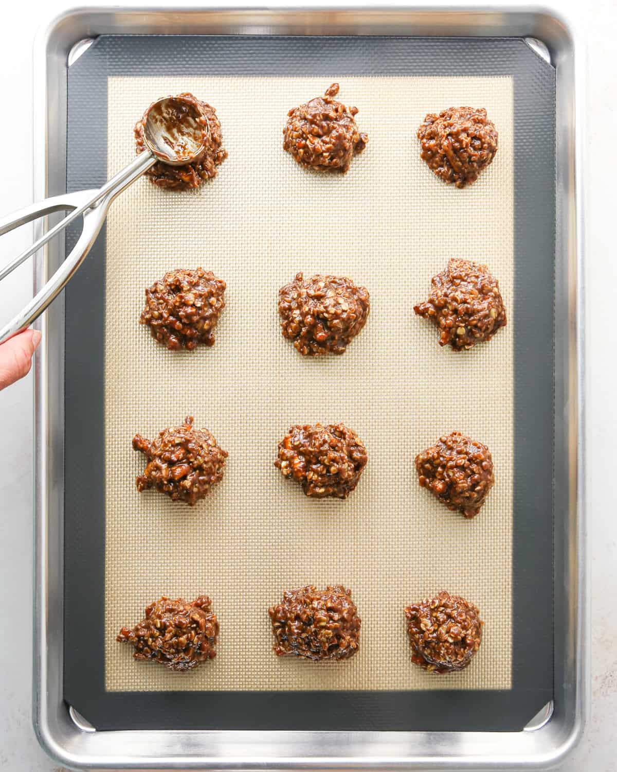 Making bird's nest cookies: overhead photo of 12 balls of cookie dough scooped onto a metal baking sheet and being pressed down with a cookie dough scoop.