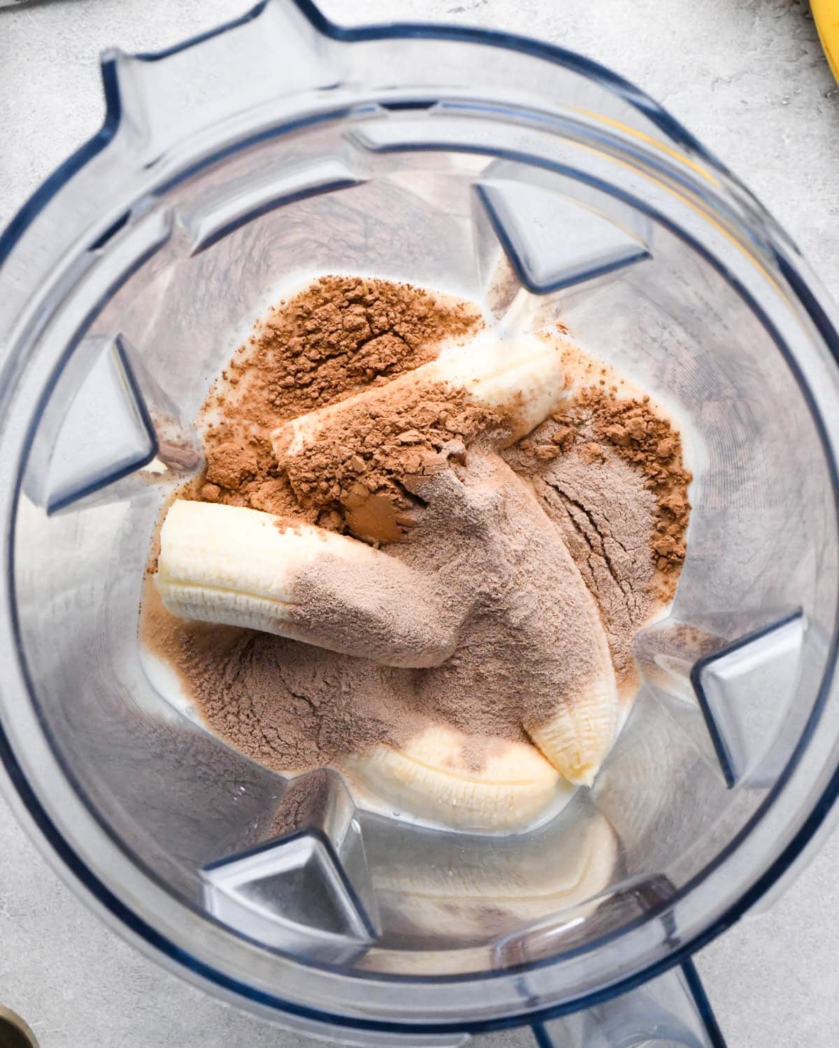 making chocolate banana smoothie: overhead photo of bananas, milk, chocolate protein powder, and cocoa powder in a blender.