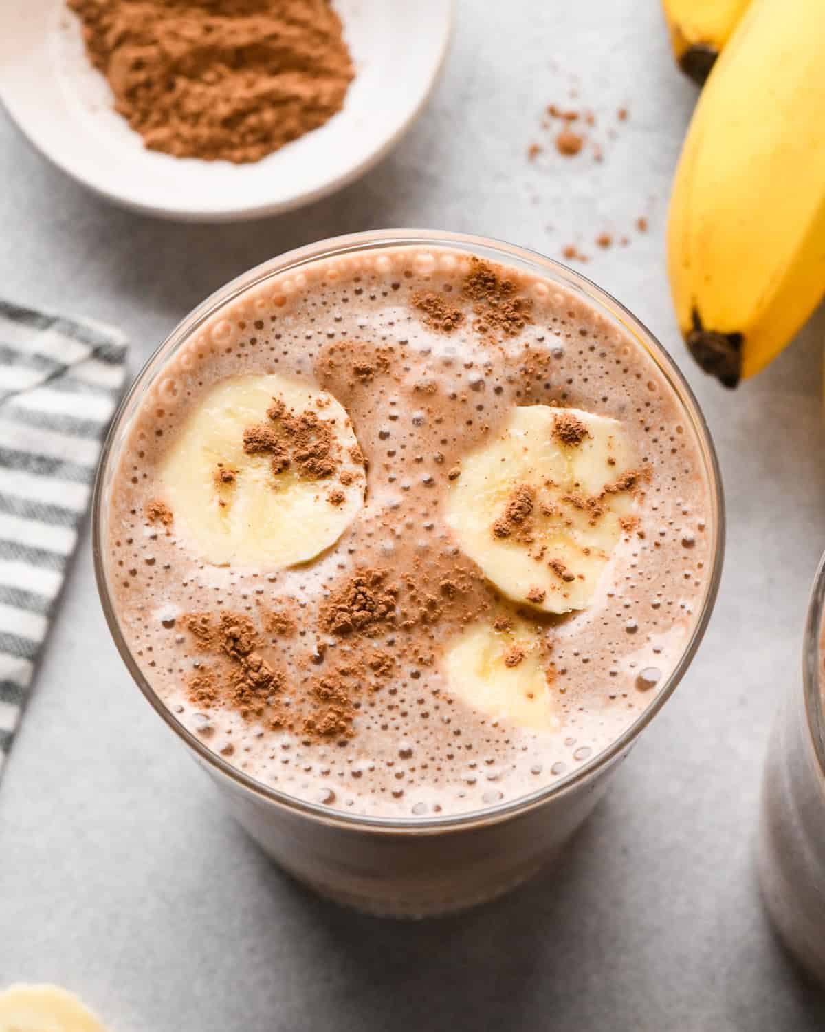 overhead photo of a chocolate banana smoothie in a clear glass topped with banana slices and cocoa powder.