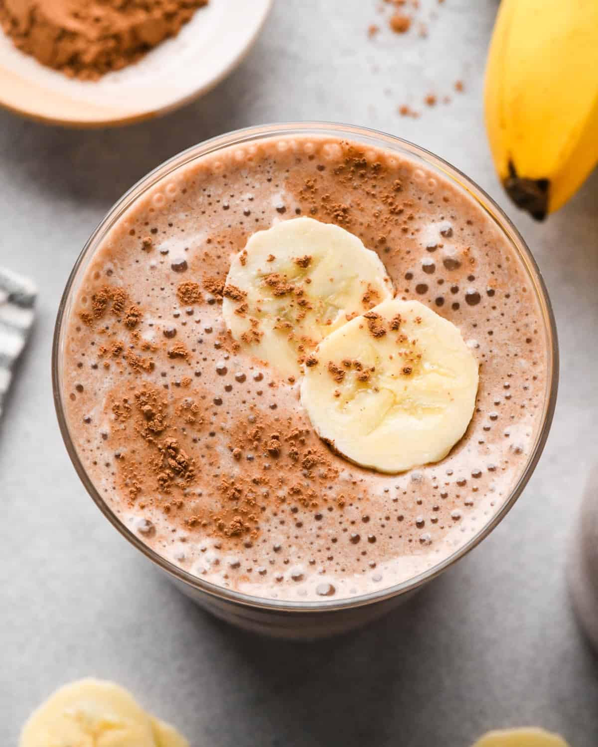 overhead photo of a chocolate banana smoothie in a clear glass topped with banana slices and cocoa powder.