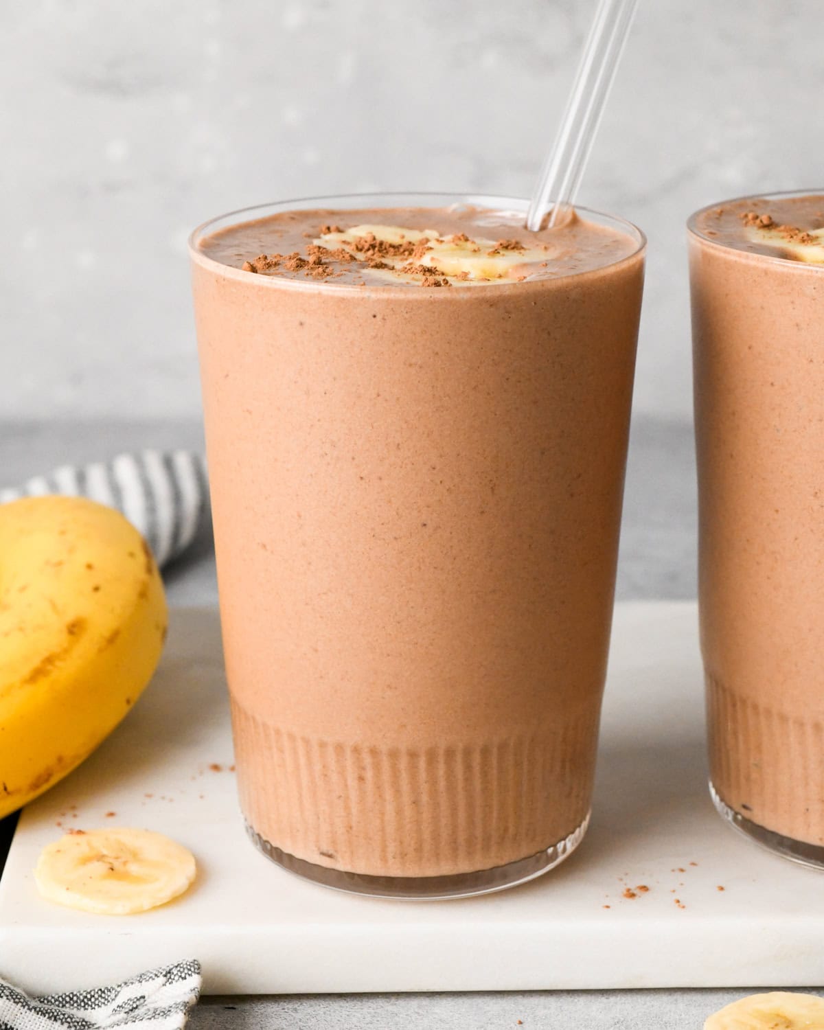 A tall glass of creamy chocolate banana smoothie topped with banana slices and cocoa powder, with a clear straw, fresh bananas, and an additional glass sitting nearby on a surface.